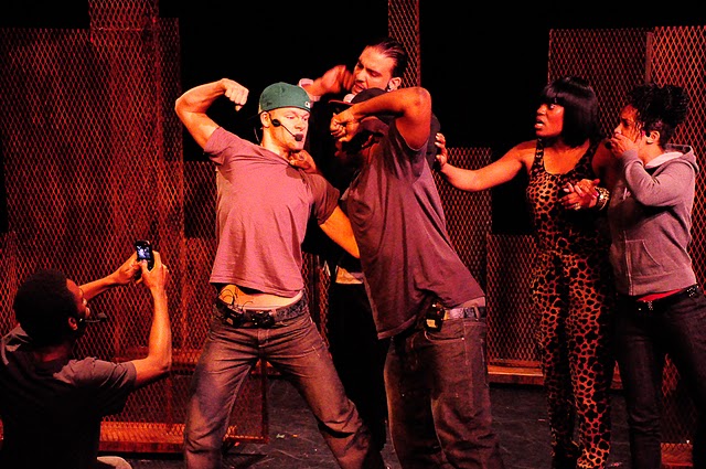 A group of young adults performing on-stage. Two people stand with their fists raised up, poised to punch each other. Another person is seated and taking a photo of them. Three other people are looking at the fight. 