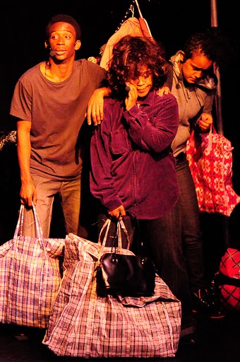 Three people performing on-stage. One person is holding a purse and a large bag in one hand and touching her other hand to their cheek. Two people stand behind them, one of which also holds a large bag.