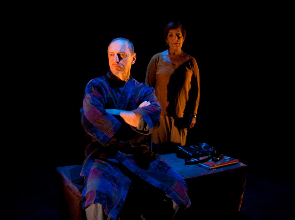 A person is performing on stage, sitting on a table facing away from another person. The seated person has their arms crossed and is looking off to the side.