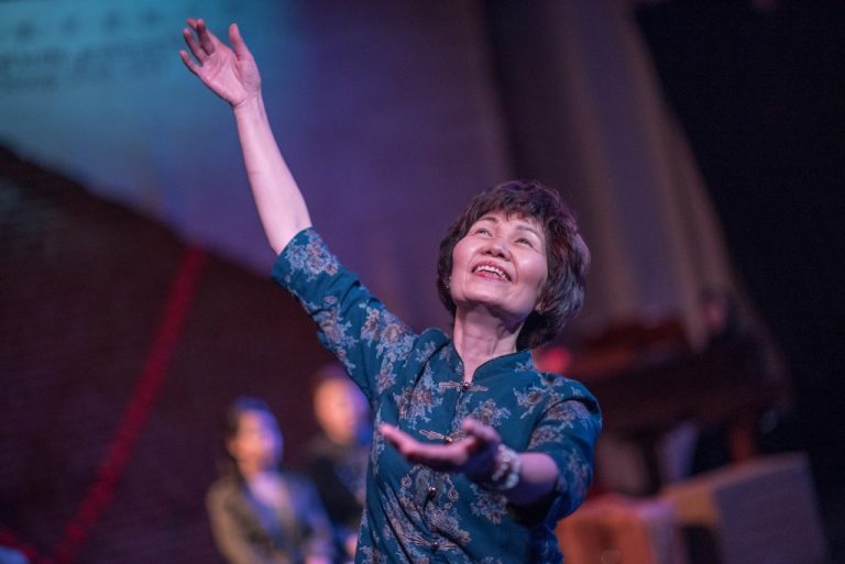 An older adult smiling with her left arm raised up and her right arm beckoning forward, in a performance of "Spring Moon".