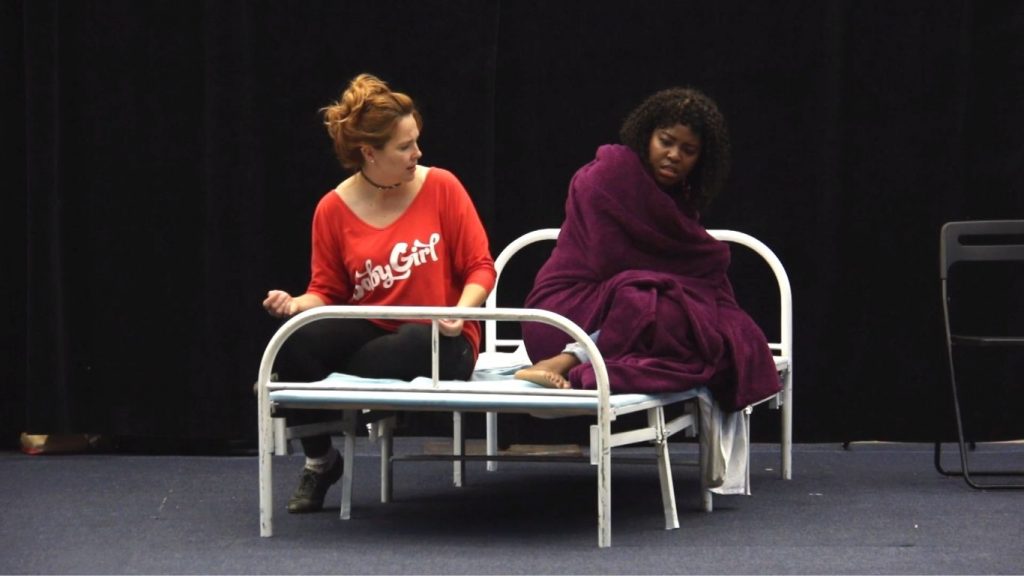 Two people performing in a post-secondary play. They are seated on a bed frame. One person is looking at the other person.