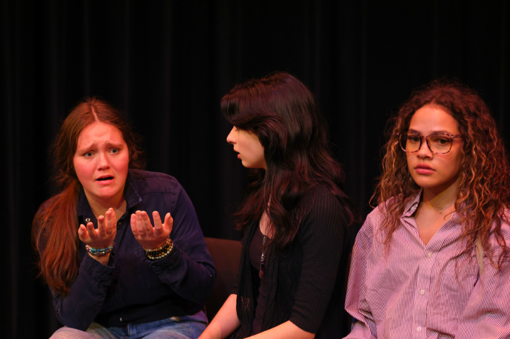 Three young people performing in a theatre production. They are all seated and the person on the left has their hands in front of them turned upwards.