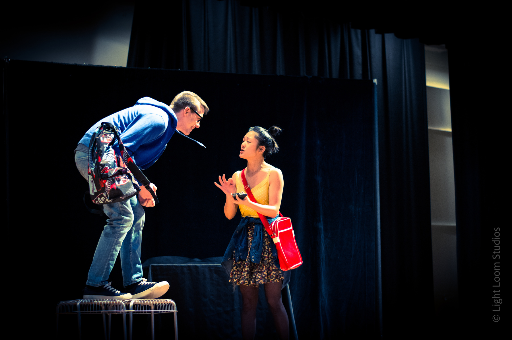 Two people performing in a theatre production onstage. One person is on top of a table leaning forward toward the person who is standing on the ground.